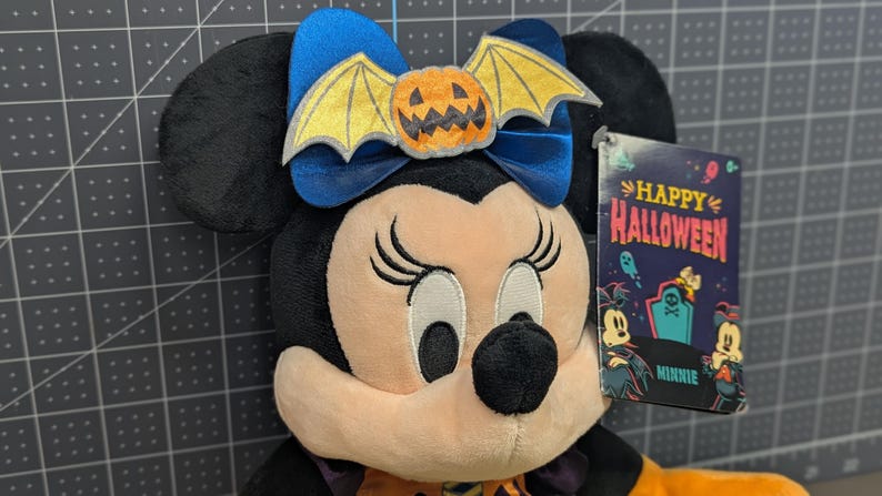 May include: A plush Minnie Mouse doll with a Halloween theme. She has black ears, a blue bow with a pumpkin bat, and a tag that says "Happy Halloween." The doll is wearing an orange and purple outfit.