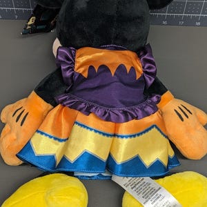 May include: A plush Minnie Mouse doll, viewed from the back, wearing a Halloween-themed dress. The dress is orange, purple, yellow, and blue. The doll has black ears, orange hands, and yellow feet with "Disney 2023" embroidered on one foot.