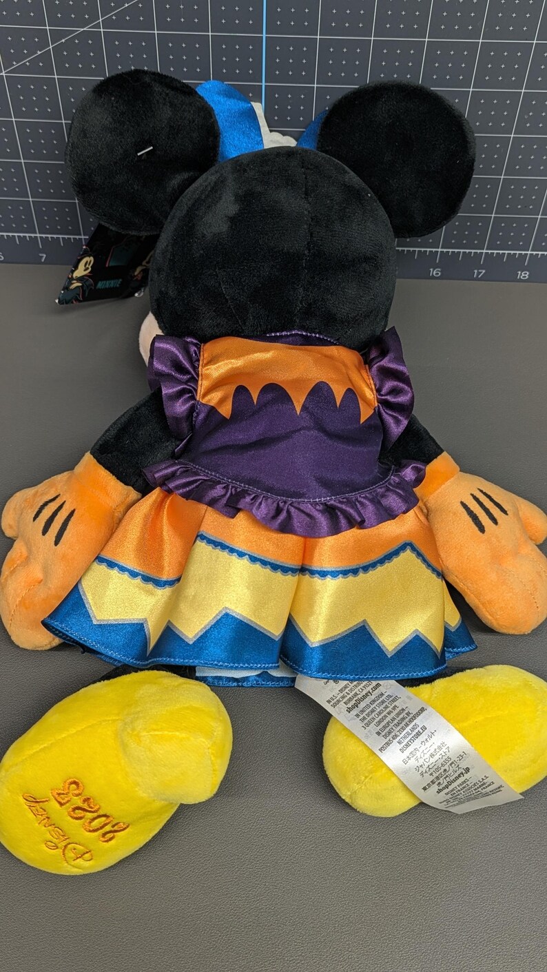 May include: A plush Minnie Mouse doll, viewed from the back, wearing a Halloween-themed dress. The dress is orange, purple, yellow, and blue. The doll has black ears, orange hands, and yellow feet with "Disney 2023" embroidered on one foot.