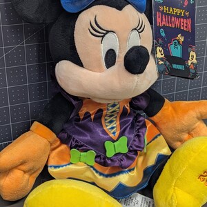 May include: A Minnie Mouse plush toy dressed in a Halloween costume. The plush has black ears, a blue bow with a pumpkin and bat design, and an orange and purple dress. A tag reads "Happy Halloween".
