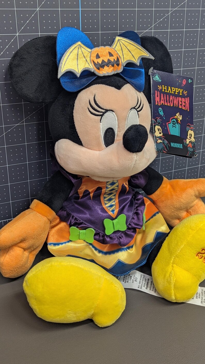 May include: A Minnie Mouse plush toy dressed in a Halloween costume. The plush has black ears, a blue bow with a pumpkin and bat design, and an orange and purple dress. A tag reads "Happy Halloween".