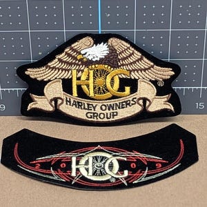 May include: Two black embroidered patches. The top patch features a gold eagle with the letters "HOG" and the words "HARLEY OWNERS GROUP". The bottom patch has the letters "HOG" with red and white accents.