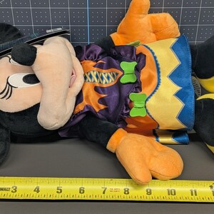 May include: A plush Minnie Mouse Halloween doll. The doll is black with a beige face, orange hands, and yellow feet. It wears a purple and orange costume with green bow accents and a bat-shaped headpiece. A measuring tape is next to the doll.