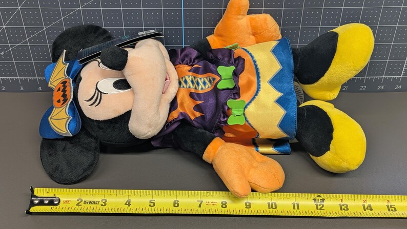 May include: A plush Minnie Mouse Halloween doll. The doll is black with a beige face, orange hands, and yellow feet. It wears a purple and orange costume with green bow accents and a bat-shaped headpiece. A measuring tape is next to the doll.