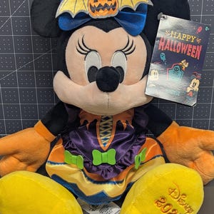 May include: A Minnie Mouse plush toy dressed in a Halloween witch costume. The plush has a blue bow with a pumpkin and bat wings, orange gloves, and yellow shoes. The tag reads "Happy Halloween" and "Disney 2023".