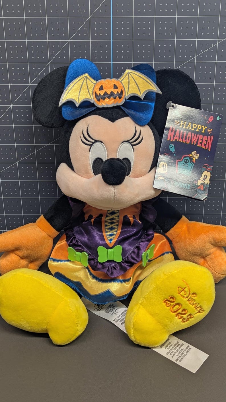 May include: A Minnie Mouse plush toy dressed in a Halloween witch costume. The plush has a blue bow with a pumpkin and bat wings, orange gloves, and yellow shoes. The tag reads "Happy Halloween" and "Disney 2023".
