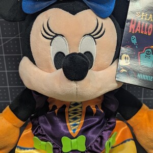 May include: A Minnie Mouse plush toy in a Halloween costume. The plush has black ears, a blue bow with a pumpkin and bat wings, and a purple and orange dress. The toy has a tag that says "Happy Halloween".