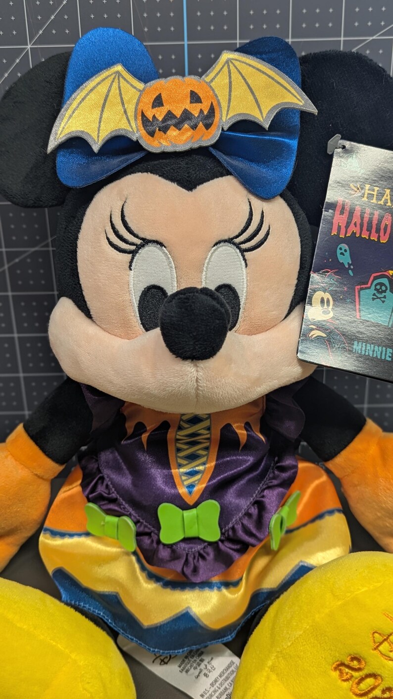 May include: A Minnie Mouse plush toy in a Halloween costume. The plush has black ears, a blue bow with a pumpkin and bat wings, and a purple and orange dress. The toy has a tag that says "Happy Halloween".