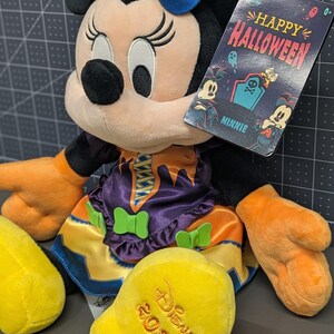May include: A Minnie Mouse plush toy dressed in a Halloween costume. The plush has a blue bow with a pumpkin and bat wings, an orange and purple dress, and orange gloves. The tag reads "Happy Halloween" and "Minnie".
