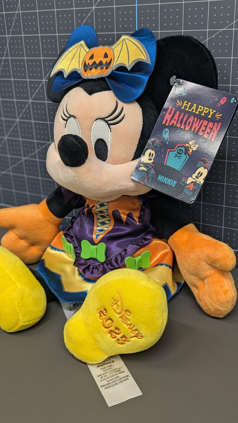May include: A Minnie Mouse plush toy dressed in a Halloween costume. The plush has a blue bow with a pumpkin and bat wings, an orange and purple dress, and orange gloves. The tag reads "Happy Halloween" and "Minnie".