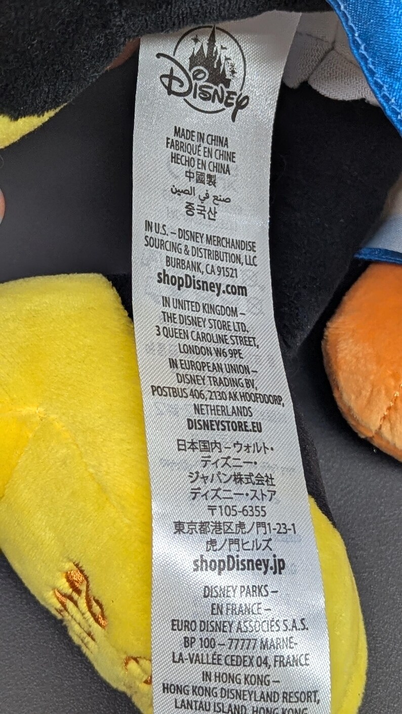 May include: A white tag with the Disney logo and text in multiple languages, including English, French, and Chinese. The tag provides sourcing and distribution information for Disney merchandise, including shopDisney.com.