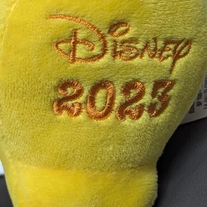 May include: Close-up of a soft, yellow plush toy with the word "Disney" and the year "2023" embroidered in a warm, orange thread. The plush has a smooth, velvety texture.