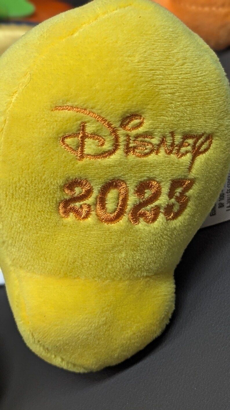 May include: Close-up of a soft, yellow plush toy with the word "Disney" and the year "2023" embroidered in a warm, orange thread. The plush has a smooth, velvety texture.