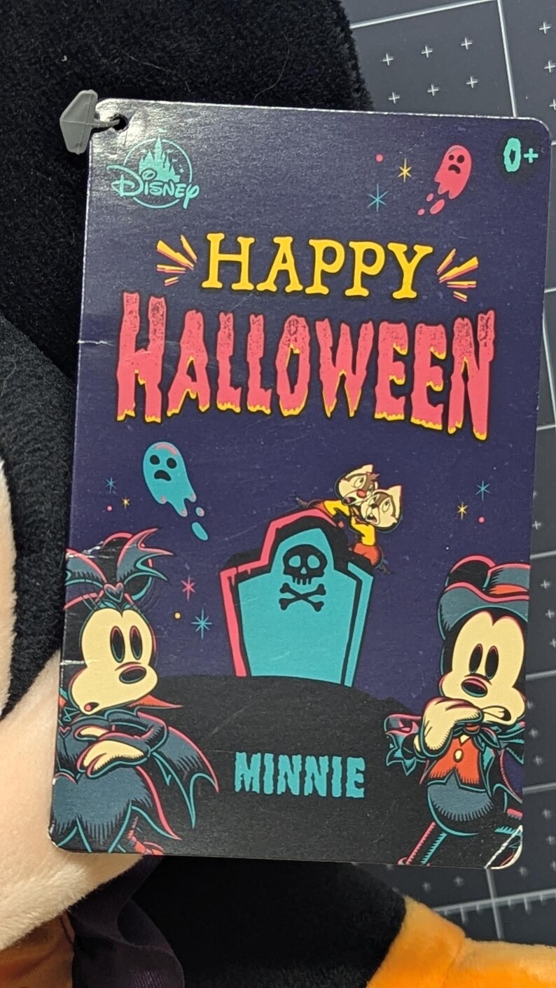 May include: A Disney Halloween-themed tag featuring Mickey and Minnie Mouse in costume. The tag reads "Happy Halloween" with images of ghosts, a tombstone, and Chip and Dale. The tag is attached to a plush toy.