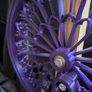May include: Close-up of a vibrant purple, gothic-style wheel with intricate, repeating arched patterns. The wheel features a central hub with a silver bolt and is likely a decorative or functional component.