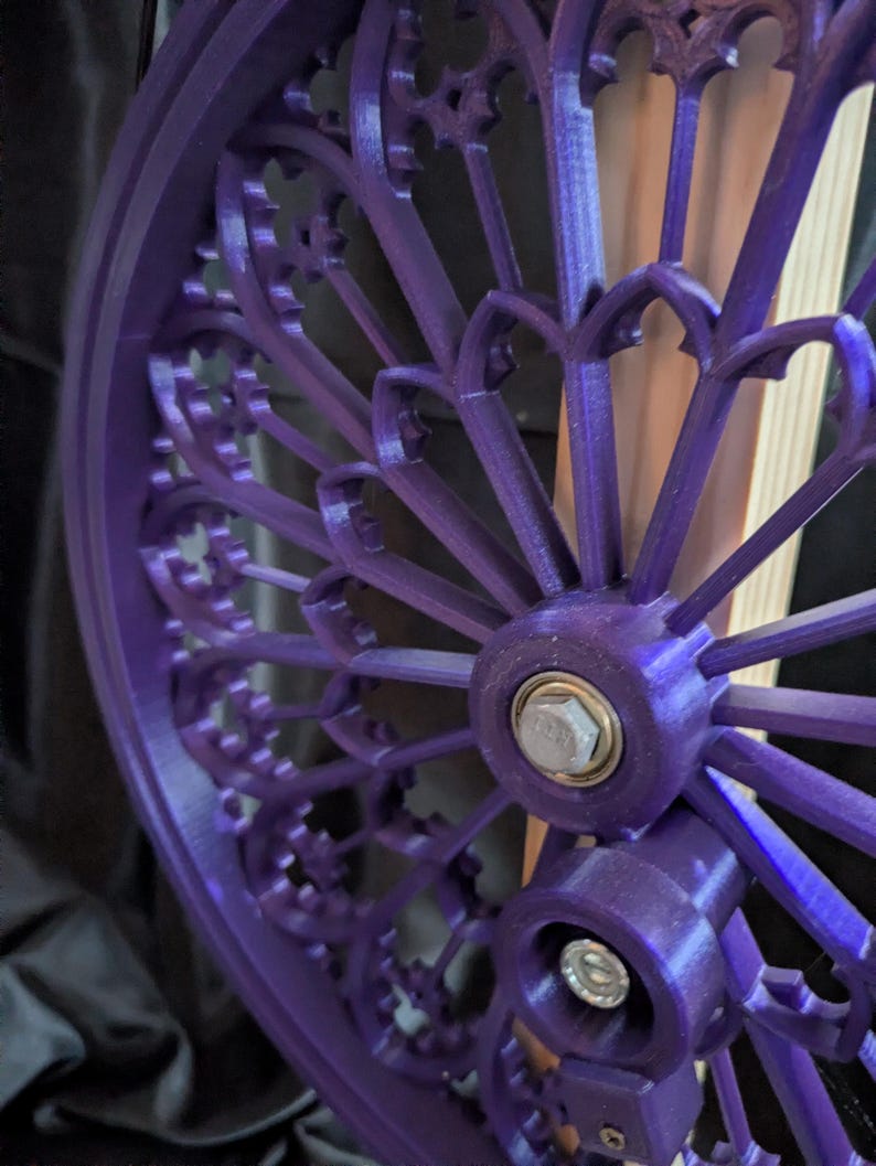 May include: Close-up of a vibrant purple, gothic-style wheel with intricate, repeating arched patterns. The wheel features a central hub with a silver bolt and is likely a decorative or functional component.