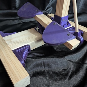 May include: A partially assembled wooden easel with purple plastic supports. The easel features light-colored wooden beams and purple, triangular-shaped supports held together with screws. The background is a black, textured fabric.