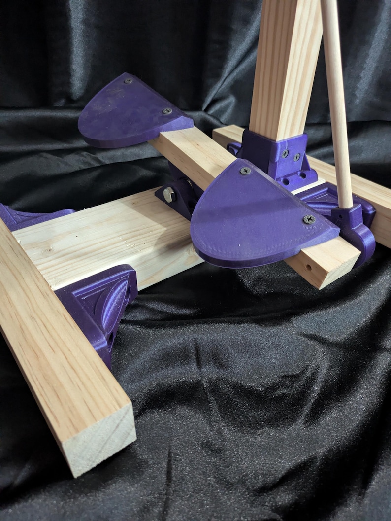 May include: A partially assembled wooden easel with purple plastic supports. The easel features light-colored wooden beams and purple, triangular-shaped supports held together with screws. The background is a black, textured fabric.
