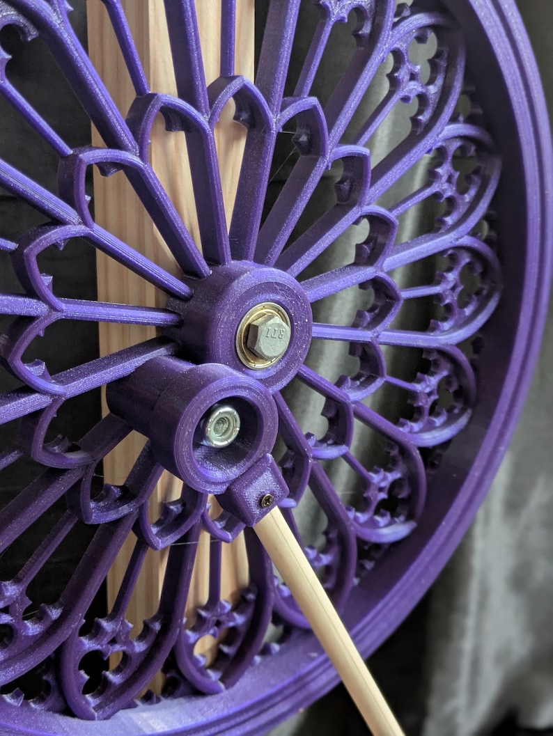May include: A close-up of a purple, decorative, circular object with an intricate, floral-like design. The object is attached to a light-colored wooden dowel with a metal bolt and nut. The design is reminiscent of a stained-glass window.