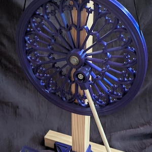 May include: A deep blue, ornate wheel with a decorative pattern, mounted on a wooden frame. The wheel is supported by a light-colored wooden dowel and a wooden base. The design is reminiscent of stained glass windows.