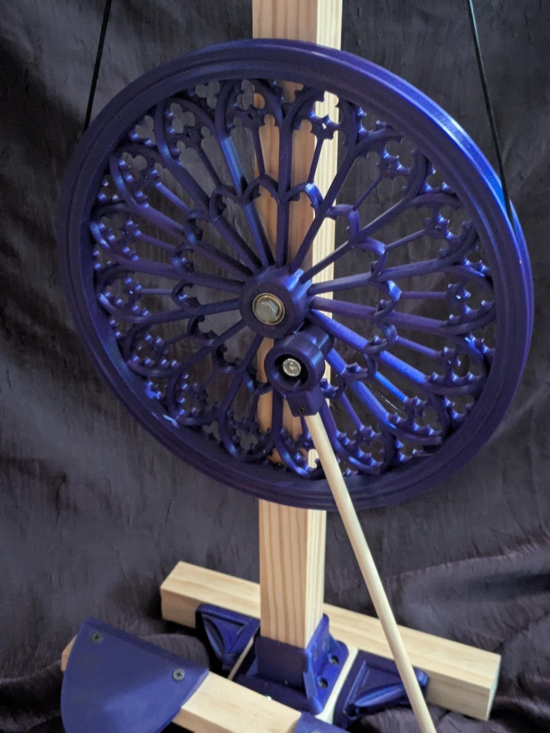 May include: A deep blue, ornate wheel with a decorative pattern, mounted on a wooden frame. The wheel is supported by a light-colored wooden dowel and a wooden base. The design is reminiscent of stained glass windows.