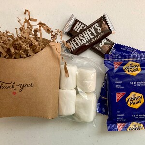 May include: A brown paper bag with the words "Thank you" and a red heart printed on it. The bag is filled with brown crinkle paper. Next to the bag are two chocolate bars, a bag of marshmallows, and two packages of Honey Maid graham crackers. The chocolate bars are Hershey's milk chocolate. The graham cracker packages are blue with a yellow Honey Maid logo.