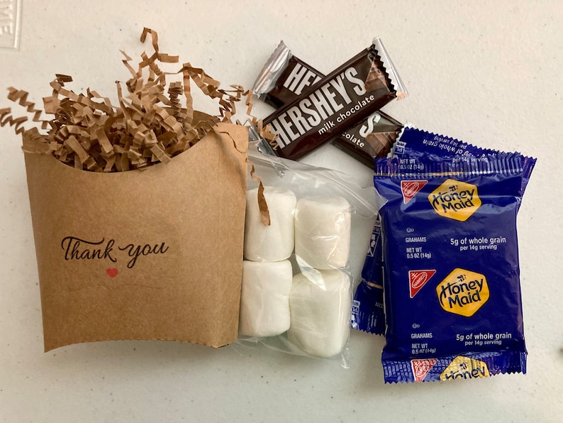 May include: A brown paper bag with the words "Thank you" and a red heart printed on it. The bag is filled with brown crinkle paper. Next to the bag are two chocolate bars, a bag of marshmallows, and two packages of Honey Maid graham crackers. The chocolate bars are Hershey's milk chocolate. The graham cracker packages are blue with a yellow Honey Maid logo.