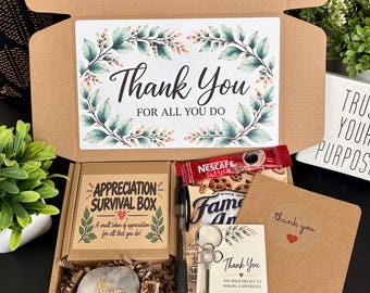 Pode incluir: Uma caixa de presente de papelão marrom contendo um cartão Appreciation Survival Box, uma pedra cinza com a inscrição You Rock, uma caneta preta, um sachê de café Nescafé, um pacote de biscoitos Famous Amos, um chaveiro em forma de chave e um cartão de agradecimento.