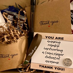 May include: A brown gift box filled with a variety of items, including chocolate bars, marshmallows, a pen, a keychain with a brown strap, and a card that reads "You are amazing, reliable, hardworking, a team player, dedicated, awesome. Thank you!"