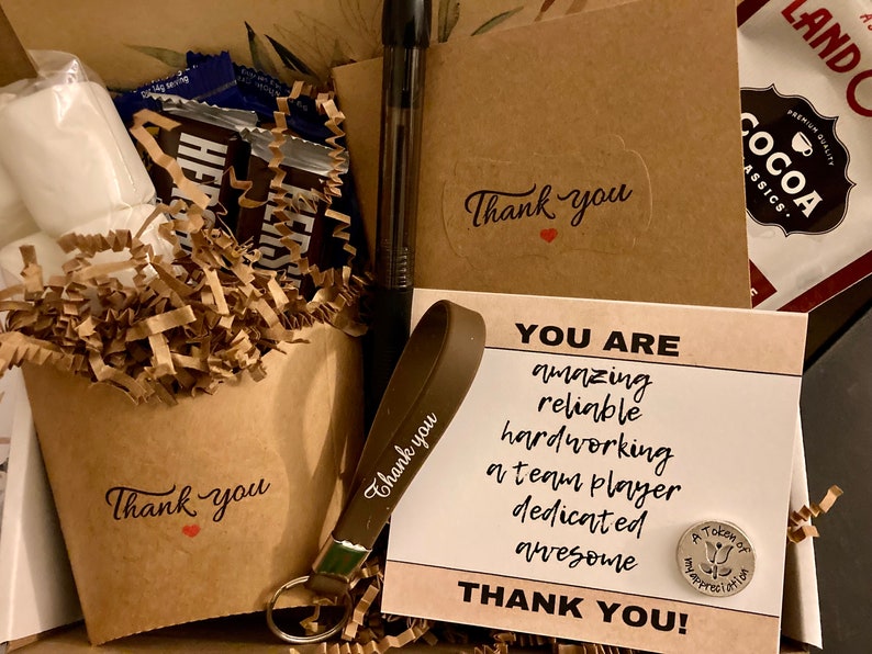 May include: A brown gift box filled with a variety of items, including chocolate bars, marshmallows, a pen, a keychain with a brown strap, and a card that reads "You are amazing, reliable, hardworking, a team player, dedicated, awesome. Thank you!"