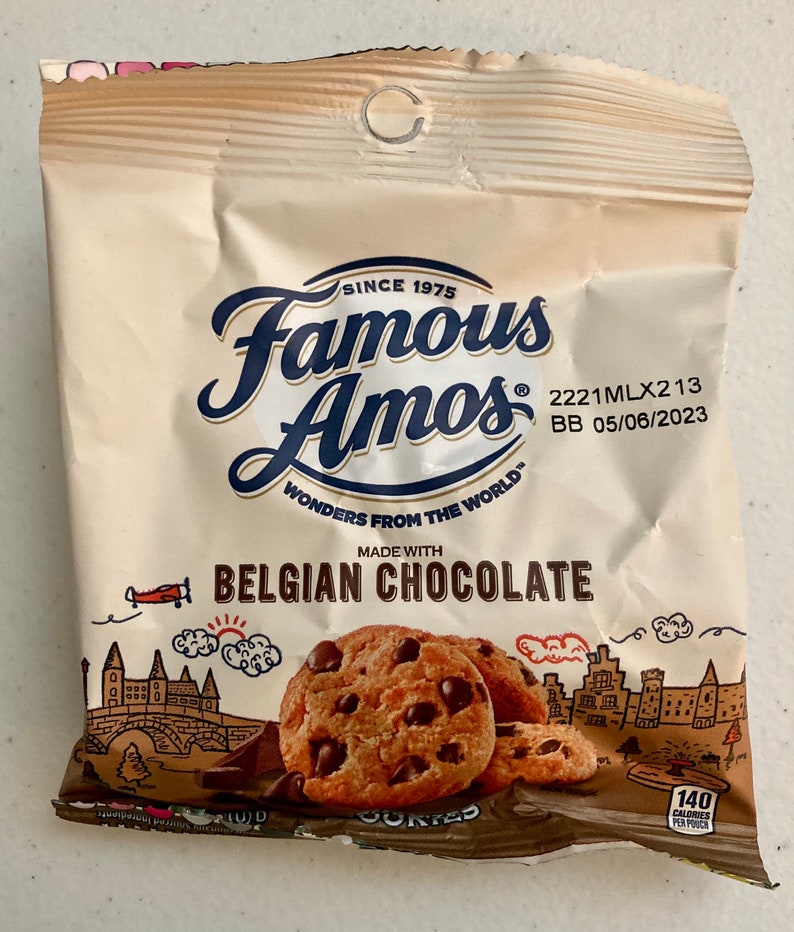 May include: A brown paper bag with a white label featuring the text "Famous Amos" and "Belgian Chocolate". The bag contains chocolate chip cookies and has a drawing of a European city with a castle and a plane.