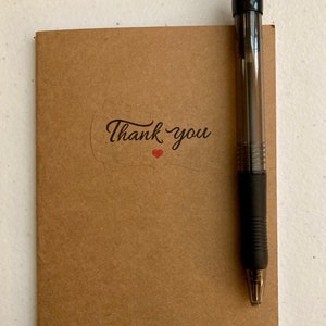 May include: A brown notebook with a thank you sticker and a red heart. A black pen is placed next to the notebook.