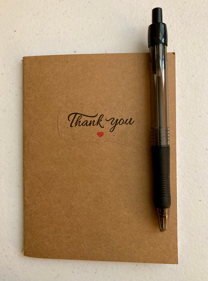 May include: A brown notebook with a thank you sticker and a red heart. A black pen is placed next to the notebook.