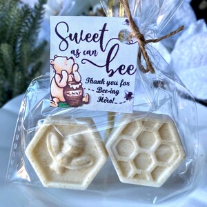 May include: Two hexagonal off-white soaps packaged in a clear plastic bag with a twine tie. One soap features a bee relief, the other a honeycomb pattern. A square tag displays a bear illustration and the text Sweet as can bee Thank you for Bee-ing Here.