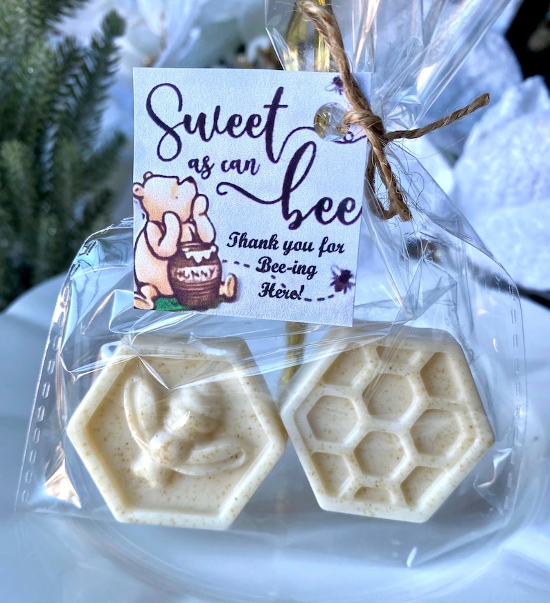 May include: Two hexagonal off-white soaps packaged in a clear plastic bag with a twine tie. One soap features a bee relief, the other a honeycomb pattern. A square tag displays a bear illustration and the text Sweet as can bee Thank you for Bee-ing Here.