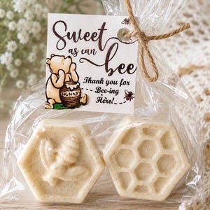 May include: Two hexagonal off-white soaps packaged in a clear plastic bag tied with twine. One soap features a bee relief, the other a honeycomb pattern. A square tag displays a Winnie the Pooh illustration and the text Sweet as can bee.