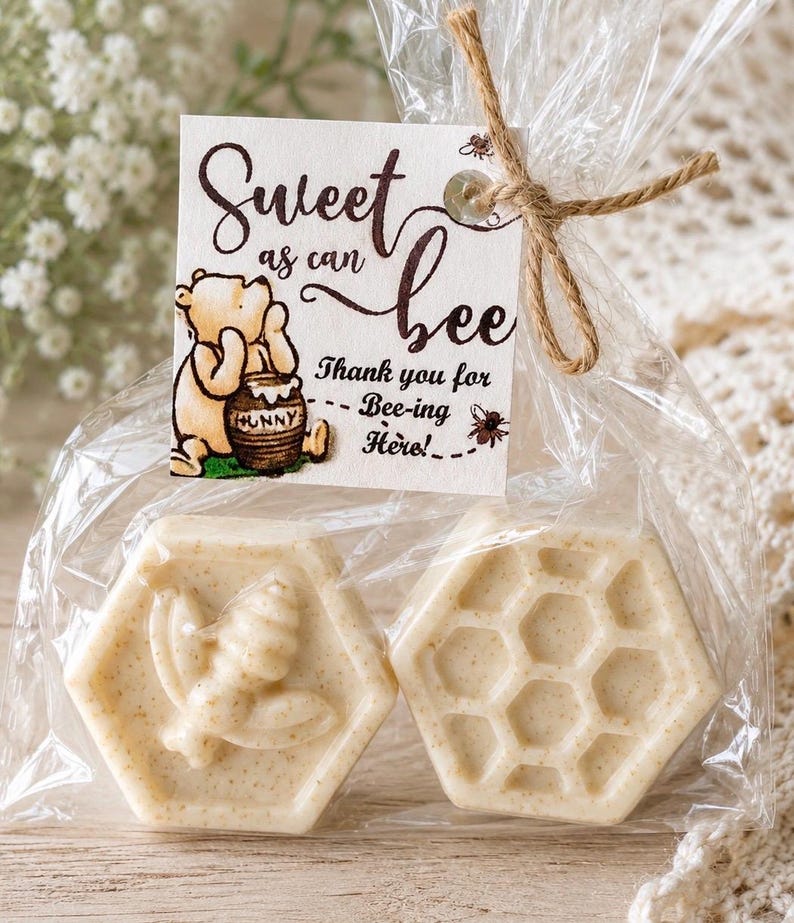 May include: Two hexagonal off-white soaps packaged in a clear plastic bag tied with twine. One soap features a bee relief, the other a honeycomb pattern. A square tag displays a Winnie the Pooh illustration and the text Sweet as can bee.