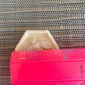 May include: A beige, hexagonal-shaped soap bar featuring a raised bee design in the center. A bright pink ruler is positioned across the front of the soap, showing a width of approximately 1.75 inches for the object.