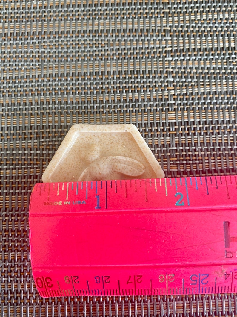 May include: A beige, hexagonal-shaped soap bar featuring a raised bee design in the center. A bright pink ruler is positioned across the front of the soap, showing a width of approximately 1.75 inches for the object.