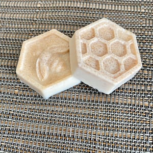 May include: Two hexagonal bars of beige soap with a speckled texture. One bar features a raised relief of a bee, while the other bar displays a raised honeycomb pattern. Both bars have a smooth, matte finish and are shaped like a hexagon.