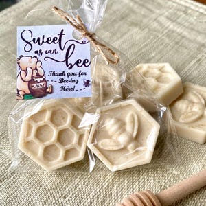 May include: A clear plastic bag containing several off-white, hexagon-shaped soaps with bee and honeycomb patterns. A square tag attached with twine features a cartoon bear, a honey pot, and the text: Sweet as can bee, Thank you for Bee-ing Here!