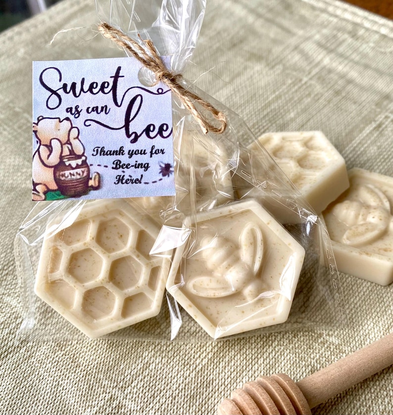 May include: A clear plastic bag containing several off-white, hexagon-shaped soaps with bee and honeycomb patterns. A square tag attached with twine features a cartoon bear, a honey pot, and the text: Sweet as can bee, Thank you for Bee-ing Here!