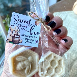 May include: Two hexagonal off-white soaps in a clear plastic bag tied with twine. One soap features a bee relief, the other a honeycomb pattern. A square tag attached to the bag shows Winnie the Pooh and the text Sweet as can bee Thank you for Bee-ing Here!
