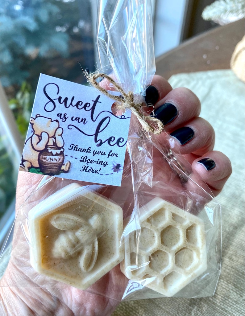 May include: Two hexagonal off-white soaps in a clear plastic bag tied with twine. One soap features a bee relief, the other a honeycomb pattern. A square tag attached to the bag shows Winnie the Pooh and the text Sweet as can bee Thank you for Bee-ing Here!