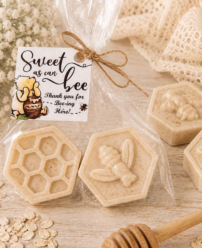 May include: Two hexagonal beige soaps in a clear plastic bag tied with twine. One soap features a honeycomb pattern, the other a bee relief. A white tag attached reads: Sweet as can bee, Thank you for Bee-ing Here!, with a Winnie the Pooh illustration.