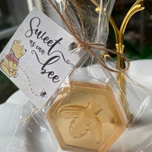 Może przedstawiać: Mały, przezroczysty woreczek foliowy wypełniony złotym mydłem w kształcie plastra miodu z wzorem pszczoły. Biała etykieta z tekstem "Sweet as can bee" jest przywiązana do woreczka sznurkiem.