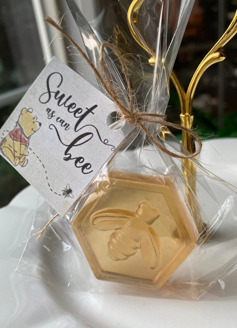 Może przedstawiać: Mały, przezroczysty woreczek foliowy wypełniony złotym mydłem w kształcie plastra miodu z wzorem pszczoły. Biała etykieta z tekstem "Sweet as can bee" jest przywiązana do woreczka sznurkiem.