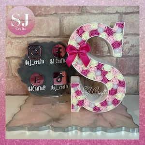 May include: A white letter S decorated with pink and cream fabric roses. The letter is on a pink and white marbled surface with a black background. The social media handles @sj_crafts, SJ Crafts, and SJCrafts19 are visible on the background.
