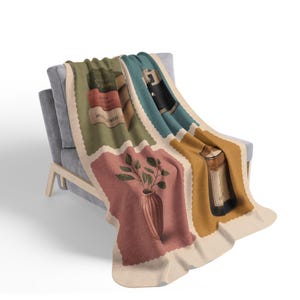May include: A soft throw blanket featuring a grid of four distinct rectangular panels. The panels display illustrations of stacked books, a black camera, a terracotta vase with green leaves, and a brown glass bottle, all on a cream background.