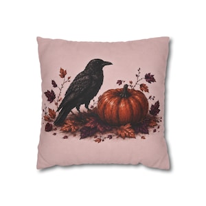Peut inclure: Un coussin carré sur fond rose clair présentant une illustration centrale d'un corbeau noir perché à côté d'une citrouille orange ronde, entouré d'un amas de feuilles d'automne rouge foncé, brunes et violettes et de fines branches.
